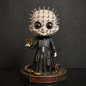 May include: A stylized figurine of a pale character with pins protruding from the head, wearing a black textured outfit with red accents. The figure holds a small gold cube and stands on a circular base with the red text HELLRAISER on the front.