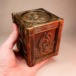 May include: A square, bronze-colored box featuring intricate relief carvings of dragons on each side and a circular dragon emblem on the top lid. The box has a metallic finish with dark shading in the recessed areas to highlight the detailed textures.