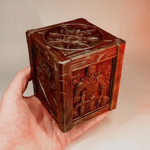 Può includere: Una scatola quadrata color rame con intricati rilievi scolpiti su ogni lato. La parte superiore mostra un emblema a forma di stella, mentre il pannello laterale raffigura una figura in armatura dettagliata che impugna una spada.