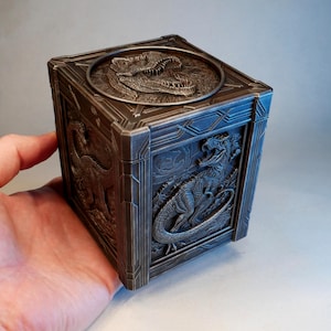 Puede incluir: Una caja cúbica de metal oscuro con intrincados relieves de dinosaurios en cada lado. La tapa superior muestra un emblema circular de una cabeza de dinosaurio, mientras que los paneles laterales exhiben figuras de dinosaurios con acabado.