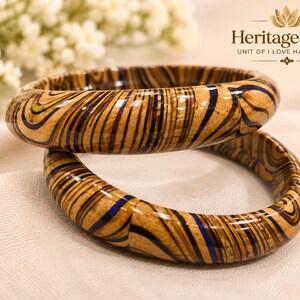 May include: Two round bangles featuring a glossy finish with swirling patterns in shades of tan, dark brown, and navy blue. The text Heritage Aura UNIT OF I LOVE HANDBLOCK is visible in the upper right corner of the image.