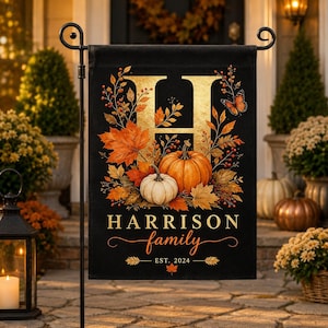 May include: A rectangular black garden flag featuring a large gold letter H, orange and white pumpkins, autumn leaves, berries, and a butterfly. The text reads HARRISON family EST. 2024 in gold lettering. The flag hangs from a black metal stand.