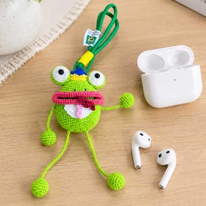 May include: A bright green crocheted frog case with a pink mouth, white belly, and blue crown detail. It features a long green cord loop for hanging and is positioned next to a white wireless earbud charging case and two white wireless earbuds.