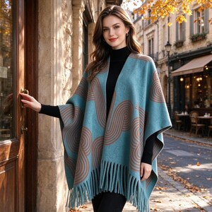 Puede incluir: Un poncho azul cerceta con un patrón geométrico marrón de líneas onduladas y un dobladillo con flecos. La prenda se lleva sobre una camiseta negra de manga larga. El poncho tiene un diseño abierto en la parte delantera y una textura suave.