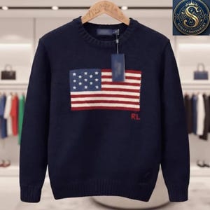 May include: A navy blue long-sleeve knit sweater featuring a prominent American flag graphic on the chest. The sweater has a ribbed crew neckline, cuffs, and hem. The letters RL are embroidered in red below the flag, and a blue tag is attached.