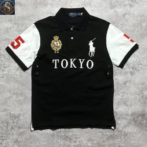 May include: A black polo shirt with white sleeves, featuring the word TOKYO in white lettering across the chest. It includes a white embroidered polo player logo, a gold crest with a Japanese flag, and the number 5 in red on the right sleeve.