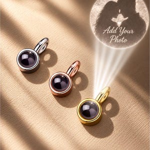 Può includere: Tre ciondoli a proiezione circolari in tonalità argento, oro rosa e oro. Ogni ciondolo presenta una pietra centrale scura e liscia. Un raggio di luce proietta la sagoma di una coppia e il testo Add Your Photo dal ciondolo dorato.