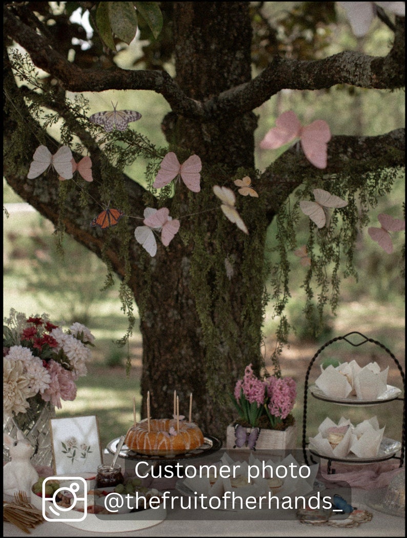 Puede incluir: Una mesa puesta con un pastel, flores y un soporte de varios niveles con vasos de papel blancos. La mesa est&aacute; decorada con una guirnalda de mariposas y un cartel que dice "Customer photo @thefruitoofherhands_"