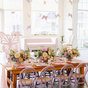 Puede incluir: Una mesa de madera con sillas moradas preparada para una fiesta infantil. La mesa est&aacute; decorada con flores rosas y blancas, y hay mariposas de papel colgando del techo. El texto "@Passthepastelevents" es visible en la esquina inferior derecha de la imagen.