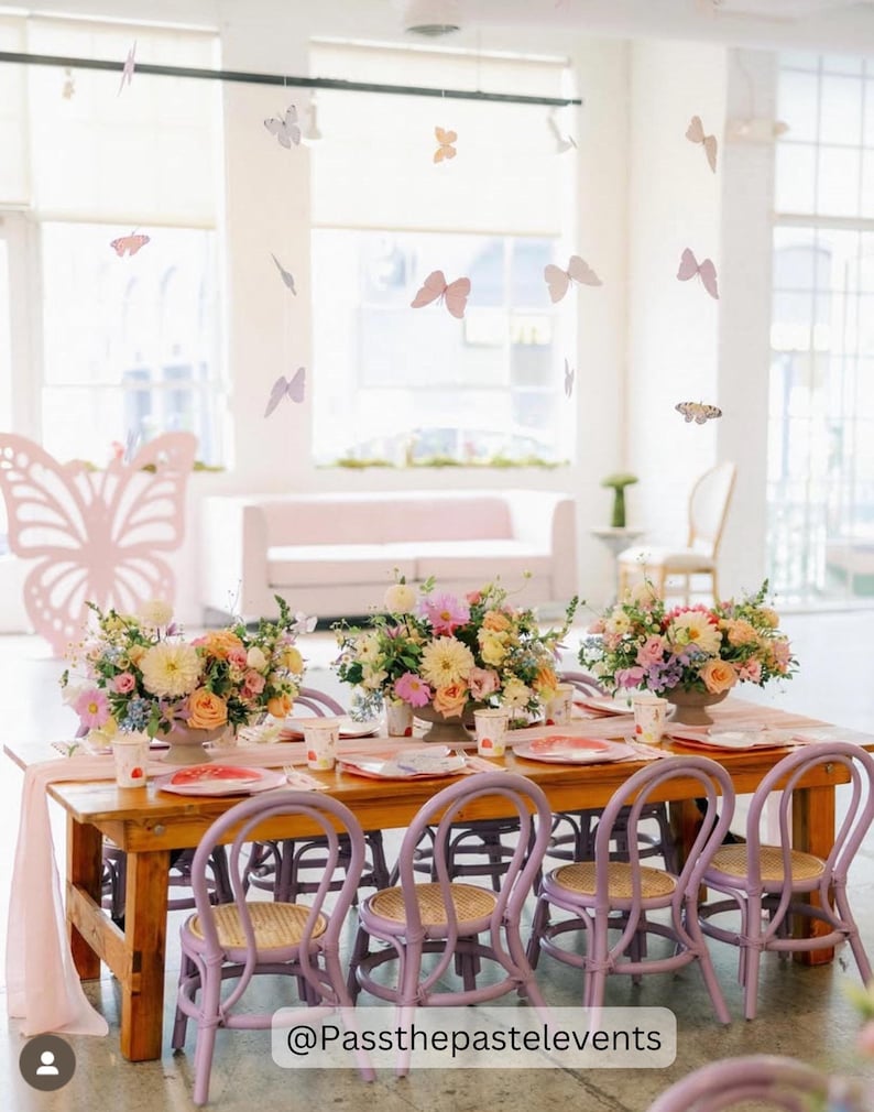 Puede incluir: Una mesa de madera con sillas moradas preparada para una fiesta infantil. La mesa est&aacute; decorada con flores rosas y blancas, y hay mariposas de papel colgando del techo. El texto "@Passthepastelevents" es visible en la esquina inferior derecha de la imagen.