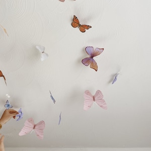 May include: A selection of colourful butterfly decorations in shades of pink, orange, purple, and blue are attached to a white ceiling. The butterflies appear to be floating, creating a whimsical and decorative effect.