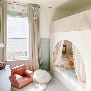 Puede incluir: Una litera blanca con un dosel sobre la litera superior. La litera inferior tiene un colch&oacute;n blanco y una almohada de margarita blanca y amarilla. La habitaci&oacute;n tiene cortinas beige, un sill&oacute;n rosa con el nombre "CORA" bordado en &eacute;l y una alfombra blanca y esponjosa. Hay mariposas colgando del techo.