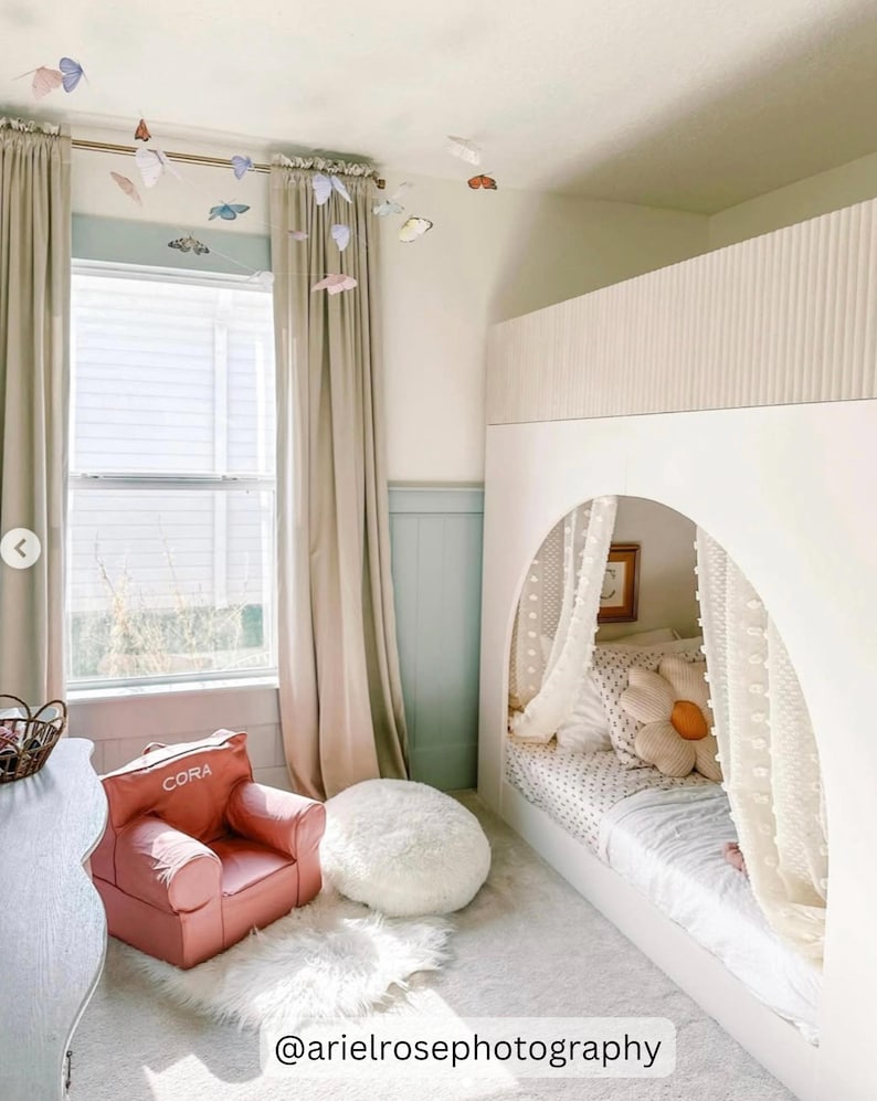 Puede incluir: Una litera blanca con un dosel sobre la litera superior. La litera inferior tiene un colch&oacute;n blanco y una almohada de margarita blanca y amarilla. La habitaci&oacute;n tiene cortinas beige, un sill&oacute;n rosa con el nombre "CORA" bordado en &eacute;l y una alfombra blanca y esponjosa. Hay mariposas colgando del techo.