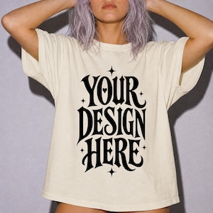 May include: A cream-colored short-sleeve t-shirt featuring bold black text that reads YOUR DESIGN HERE in a stylized font. The graphic includes small star accents and a heart shape within the letter O, centered on the front of the garment.