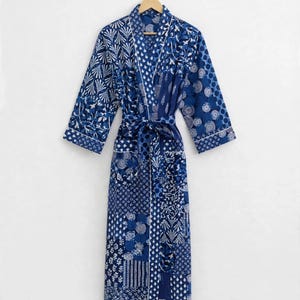May include: A long-sleeved robe featuring a patchwork design of various blue and white geometric and floral patterns. The garment includes a matching tie belt at the waist, two front pockets, and white piping along the collar and sleeve cuffs.