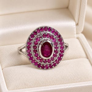 Puede incluir: Un anillo de tono plateado con un rubí central grande de corte ovalado, rodeado por dos halos concéntricos de rubíes más pequeños de corte redondo. La banda de metal tiene un diseño de brazo dividido y un acabado pulido brillante.
