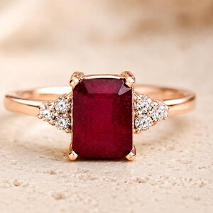 Puede incluir: Un anillo de oro rosa con una gran piedra central de rubí rectangular de color rojo intenso, flanqueada por tres pequeños diamantes redondos transparentes a cada lado. El anillo lleva el logotipo de Virasat Jewels en la esquina superior.