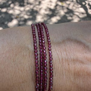 May include: A multi-strand bracelet composed of small, faceted, deep red garnet beads. The bracelet wraps around the wrist in four distinct rows, showcasing the reflective, dark crimson surfaces of the individual circular stones in a continuous line.