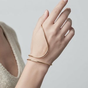 Könnte beinhalten: Ein Handarmband aus goldfarbenem Metall mit einem glatten, durchgehenden Drahtdesign, das sich um das Handgelenk legt und in einer geschwungenen, wellenartigen Form über den Handrücken verläuft, was eine schlanke und offene Struktur erzeugt.