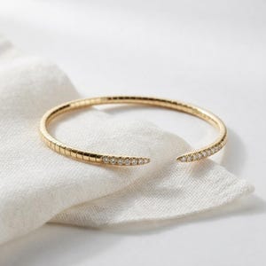 Puede incluir: Un brazalete de oro con un diseño de banda flexible y segmentada. Los dos extremos cónicos del brazalete están adornados con una hilera de pequeñas piedras redondas y transparentes, creando un acabado brillante en ambos lados de la joya abierta.