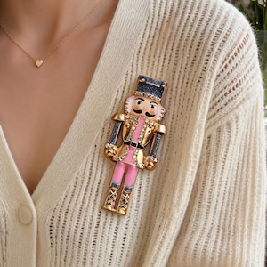 May include: A nutcracker brooch featuring a gold-tone metal finish with pink enamel accents on the jacket and trousers. The figure is adorned with clear rhinestones along the arms, legs, and hat, and includes a dark blue glittered hat top.