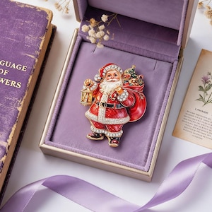 May include: A Santa Claus brooch featuring a red suit with white trim, a black belt, and gold accents. The figure holds a gold lantern in one hand and carries a large red sack filled with gifts over the shoulder. The design is embellished with clear crystals.
