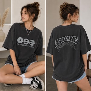 Peut inclure: Un t-shirt gris anthracite à manches courtes avec un motif frontal composé de trois icônes circulaires et du texte BTS WORLD TOUR | ARIRANG. Le dos du t-shirt présente le texte arqué ARIRANG WORLD TOUR dans une police de caractères en gras.