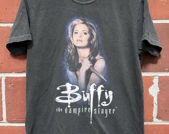 May include: A charcoal gray short-sleeve t-shirt featuring a graphic print of a woman holding a wooden stake. Below the image, the white text reads Buffy the Vampire Slayer. The shirt has a crew neckline and is displayed on a metal hanger.