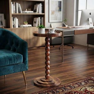 May include: A round wooden side table featuring a central pedestal composed of stacked spheres, resting on a tiered circular base. A small white ceramic potted plant and a white coffee cup on a saucer are placed on the tabletop surface.