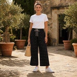 May include: A white short-sleeved crew neck t-shirt paired with black and grey gingham print wide-leg trousers featuring a drawstring waist. The outfit is completed with white sneakers and a small, light brown woven straw handbag.