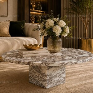 May include: A round coffee table with a white and grey marble top and a square marble base. On the table sits a ribbed glass vase filled with white hydrangea flowers, a gold-toned decorative bowl, and two stacked books titled Luxury Living.