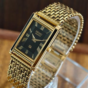 May include: A gold-tone rectangular wristwatch featuring a black dial with gold Roman numeral hour markers. The watch has a textured gold-tone metal link bracelet and displays the text SEIKO QUARTZ on the face. The watch case is polished gold.
