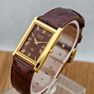 May include: A rectangular Seiko Quartz wristwatch featuring a gold-tone case, a textured brown dial with gold Roman numeral hour markers, and a brown leather strap with a crocodile-embossed pattern and a gold-tone buckle closure.