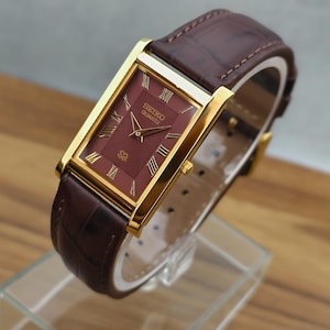 May include: A rectangular Seiko Quartz watch with a gold-tone case and a deep red dial featuring a textured pattern and gold Roman numeral hour markers. The watch is fitted with a dark brown leather strap with a visible crocodile-style texture.