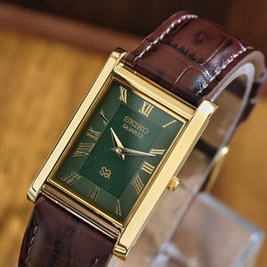 May include: A rectangular Seiko Quartz wristwatch featuring a dark green dial with gold-tone Roman numeral hour markers and hands. The watch has a polished gold-tone case and a brown leather strap with a textured, embossed crocodile-skin pattern.