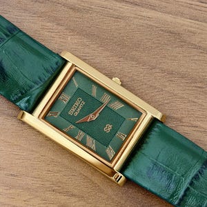 May include: A rectangular gold-tone wristwatch featuring a green dial with a textured center, gold Roman numeral hour markers, and gold hands. The watch includes a green leather strap with a crocodile-embossed pattern and the text Seiko Quartz.