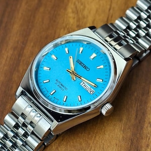 May include: A Seiko automatic wristwatch with a bright turquoise dial, silver-tone hour markers, and gold-tone hands. The watch features a day-date window at the three o'clock position, a stainless steel case, and a multi-link metal bracelet.