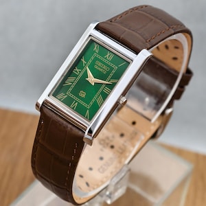 May include: A rectangular Seiko Quartz watch with a green dial, gold-tone hands, and Roman numeral hour markers. The watch features a silver-tone metal case and a brown leather strap with a crocodile-embossed texture and a buckle closure.