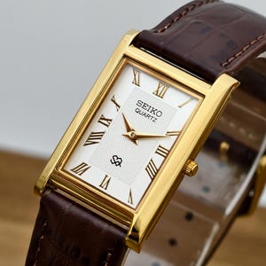 May include: A rectangular Seiko Quartz wristwatch with a gold-tone case, a white dial featuring a wavy texture, gold-tone hands, and black Roman numeral hour markers. The watch includes a dark brown leather strap with a textured crocodile-like pattern.