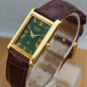 Peut inclure: Une montre-bracelet rectangulaire Seiko Quartz avec un boîtier doré, un cadran vert foncé à motif texturé et des index en chiffres romains dorés. La montre est dotée d'un bracelet en cuir marron avec une texture gaufrée façon crocodile.