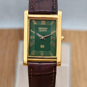 May include: A rectangular Seiko Quartz watch with a gold-tone case and a dark brown leather strap. The watch face is green with a textured center, gold-colored Roman numeral hour markers, and gold-tone hands. The text Seiko Quartz is printed.