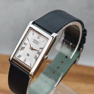 May include: A rectangular Seiko Quartz wristwatch featuring a silver-toned case, a white dial with black Roman numeral hour markers, and rose gold-toned hands. The watch includes a black leather strap with a textured finish and a light green lining.