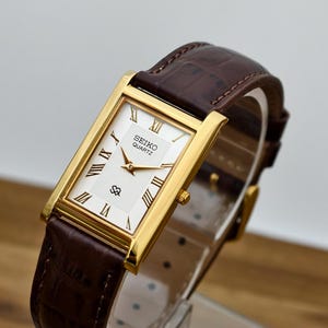 May include: A rectangular gold-tone Seiko Quartz wristwatch featuring a white dial with Roman numeral hour markers, gold hands, and a textured pattern. The watch is paired with a dark brown leather strap with a crocodile-embossed texture.