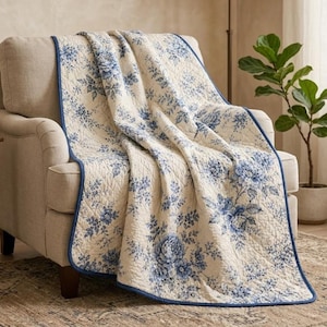 May include: A cream-coloured armchair draped with a quilted throw blanket featuring a blue floral pattern on a white background. The blanket has a solid blue fabric border and is folded over the back and seat of the upholstered chair.