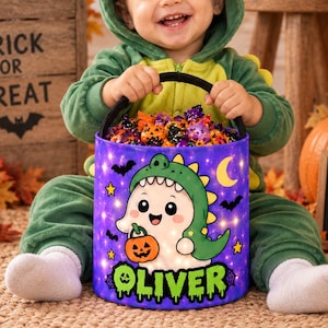 May include: A purple Halloween bucket featuring a cartoon ghost wearing a green dinosaur costume, holding a pumpkin, and the name Oliver in green text. The bucket is filled with assorted candies and is held by a child in a green dinosaur onesie.