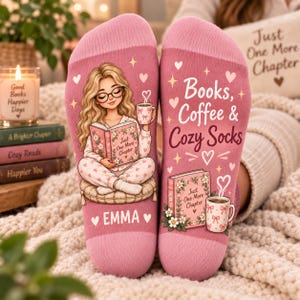 May include: A pair of pink socks featuring a graphic of a person with long blonde hair reading a book and holding a mug. The text reads Books, Coffee & Cozy Socks, Just One More Chapter, and EMMA, decorated with hearts and small stars.