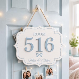 May include: A white, scalloped-edge wooden sign with light blue text reading ROOM 516 and MIA & CHLOE, featuring a small blue bow graphic. The sign is suspended by a tan rope from a white hook, displaying a clean, light-colored design for a door.