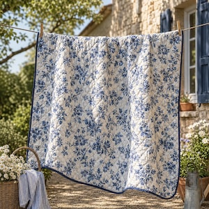 Puede incluir: Una manta acolchada rectangular con base blanca y un patrón floral y botánico azul repetitivo. La manta tiene un borde de tela azul oscuro en los cuatro lados y está suspendida de un alambre fino mediante dos pinzas de madera para ropa.
