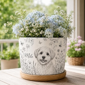 Puede incluir: Una maceta de cerámica blanca con un dibujo de línea negra de un perro y el nombre Milo, rodeado de ilustraciones botánicas de flores silvestres y mariposas. La maceta se asienta sobre una base de madera circular y contiene flores azul claro.