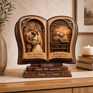 Op de afbeelding: Een bruin houten sculptuur in de vorm van een boek met twee uitgesneden scènes: een bruidspaar links met de tekst From Our Vows en een ouder echtpaar op een bankje rechts met de tekst To Our Forever. De voet toont 1976-2026, Happy 50th Anniversary en James ♡ Emma.